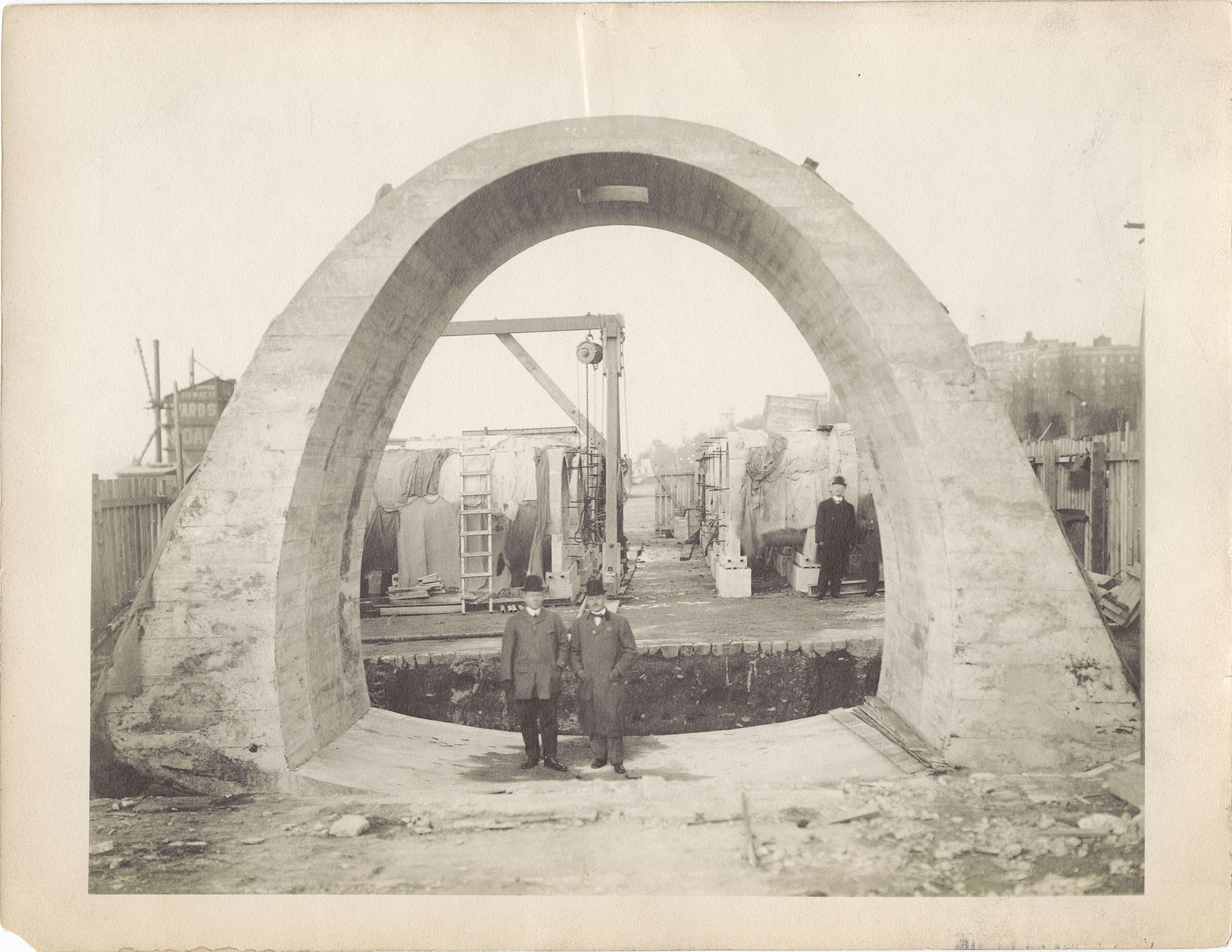 Black and white photo of two men standing under a large concrete arch about four times their height.