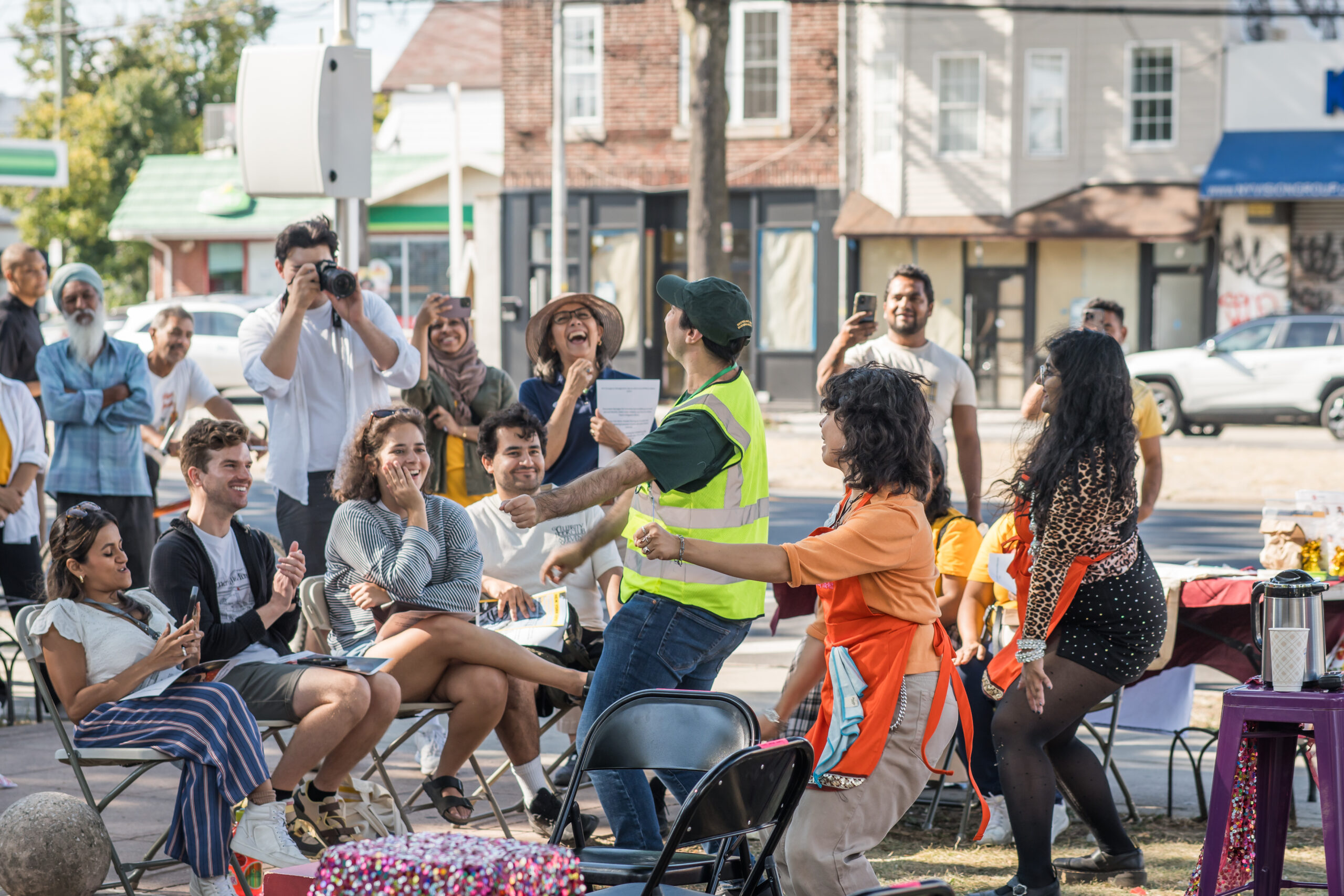 A group of performers dances as part of a play staged in public. Audienc emembers laugh, applaud and take photos either standing or in folding chairs.