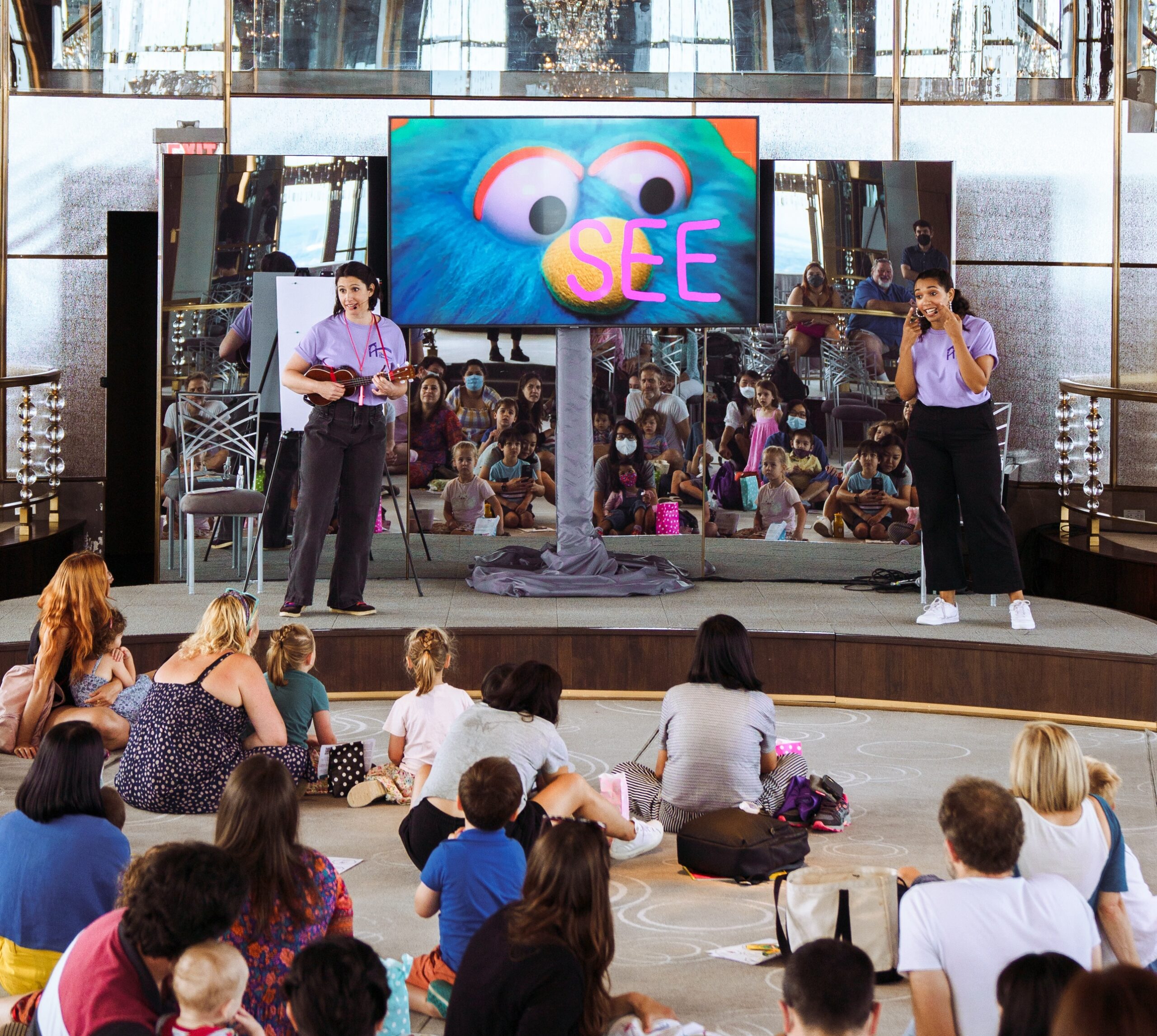 Two women in light purple t-shirts sing and play ukelele on either side of a large video screen which displays a large close up of a bright blue puppet. An audience watches.