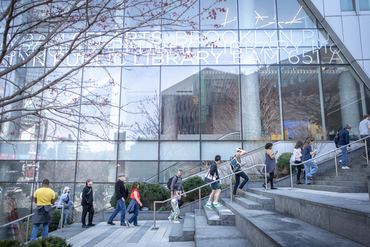 People walk up and down stairs outside a glass-walled building labeled Brooklyn Public Library.