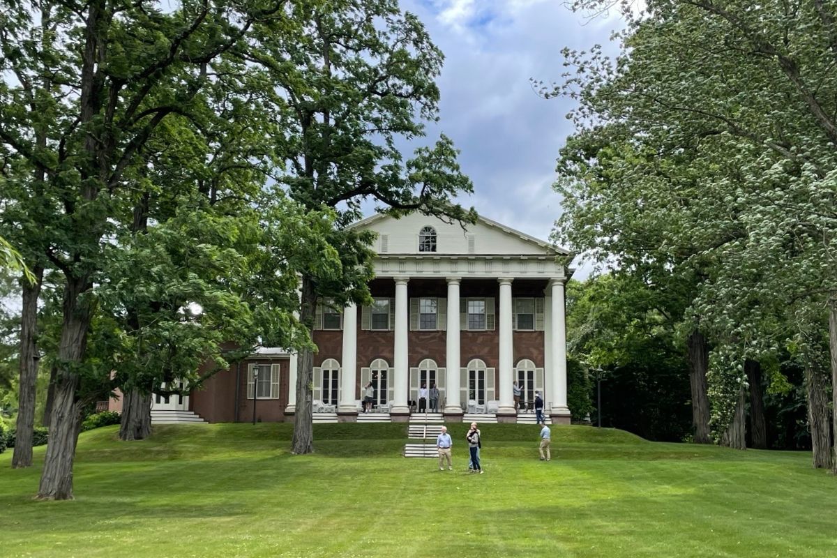 The exterior of a mansion in neoclassical style with six white columns upholding a white pediment and triangular roof. Several visitors stand on the broad porch and expanse of rolling lawn in front of the building.