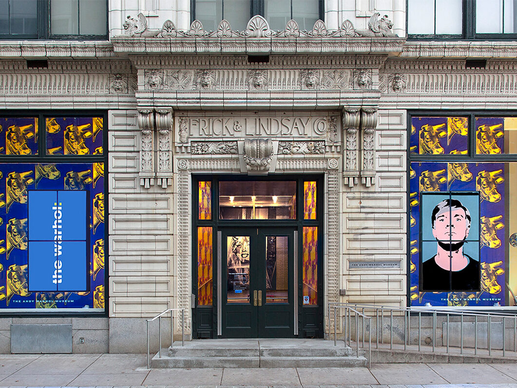 A photograph of the Andy Warhol Museum entrance. It is an ornate building with cement stairs and an accessible ramp that leads off the right side of the image. A portrait of Marilyn Monroe is visible through glass panels in and around the front doors. The front door is flanked by two large windows with blue wallpaper featuring screen prints of yellow cows. The window on the left side of the image has a sign that reads The Warhol in white print. The right window has a screen-printed portrait of Andy Warhol.