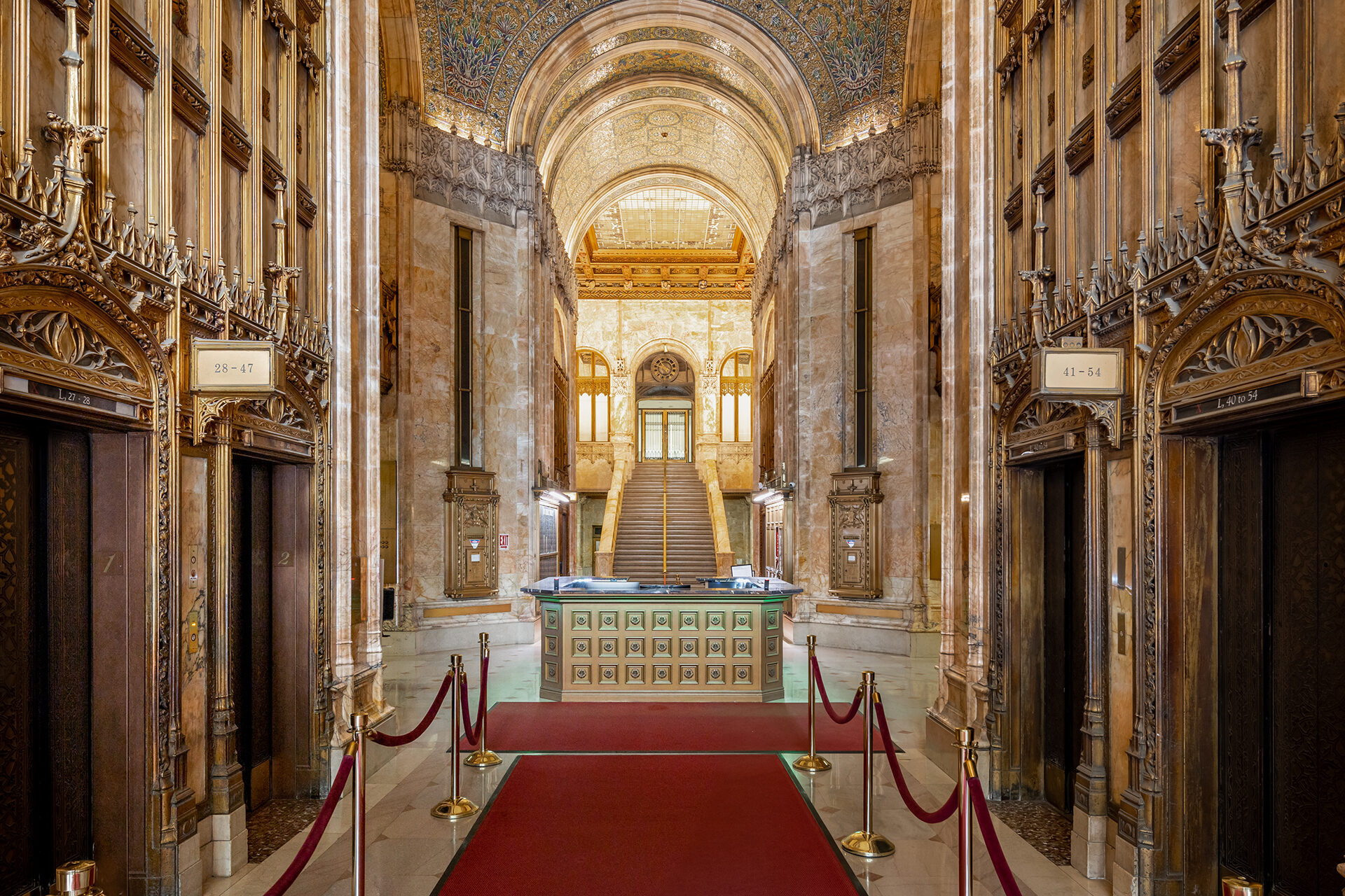 Ornate lobby with marble walls, arched ceiling, elevators, red carpet, and a grand staircase in the background.