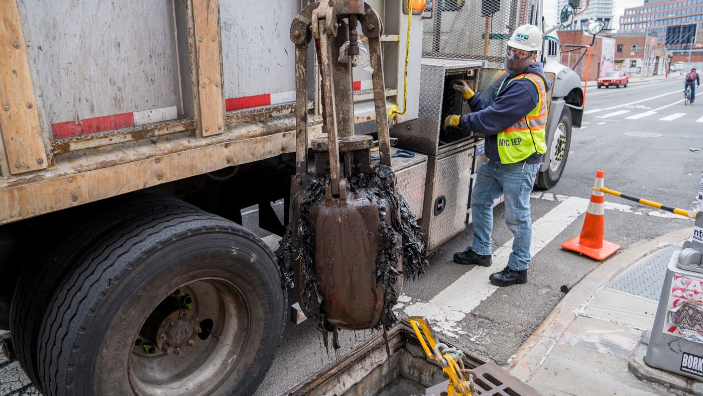 A large white dump truck with a metal claw arm that is being operated by a worker. The claw is filled with slimy black sewer detritus pulled from a catch basin standing open just below the claw.
