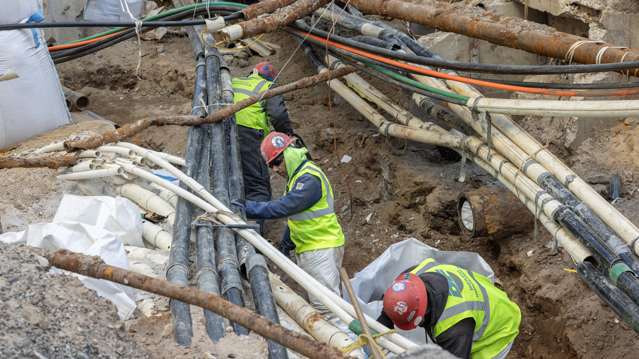 Three men in yellow vests and hard hats conduct work in a llarge trench, surrounded by dirt and a jumble of wires and pipes every which way around them.