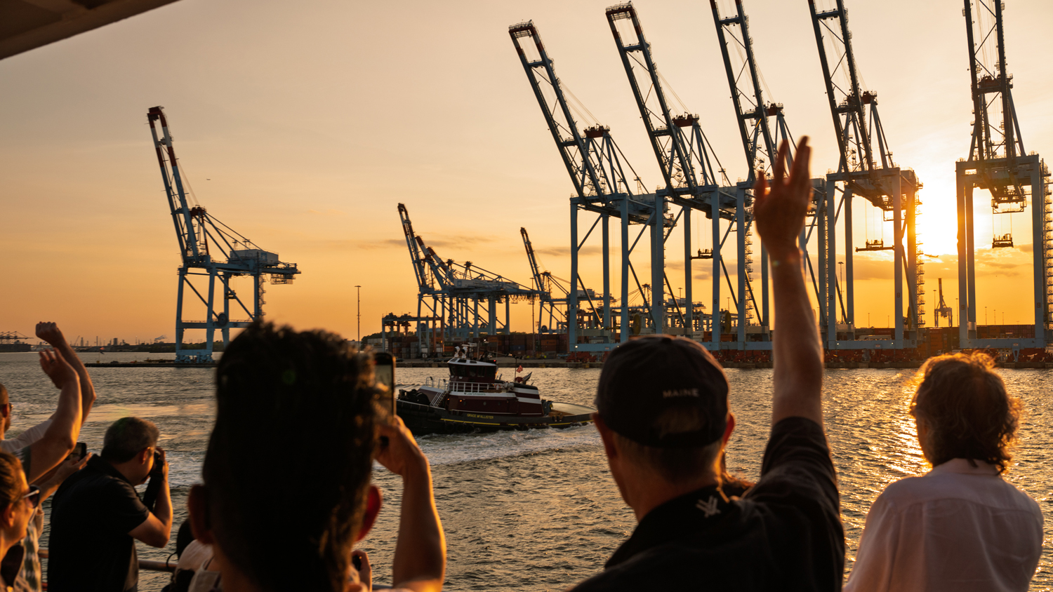 People in the foreground look out over a waterway at sunset at a tugboat and many gantry cranes silhouetted against the sky. 