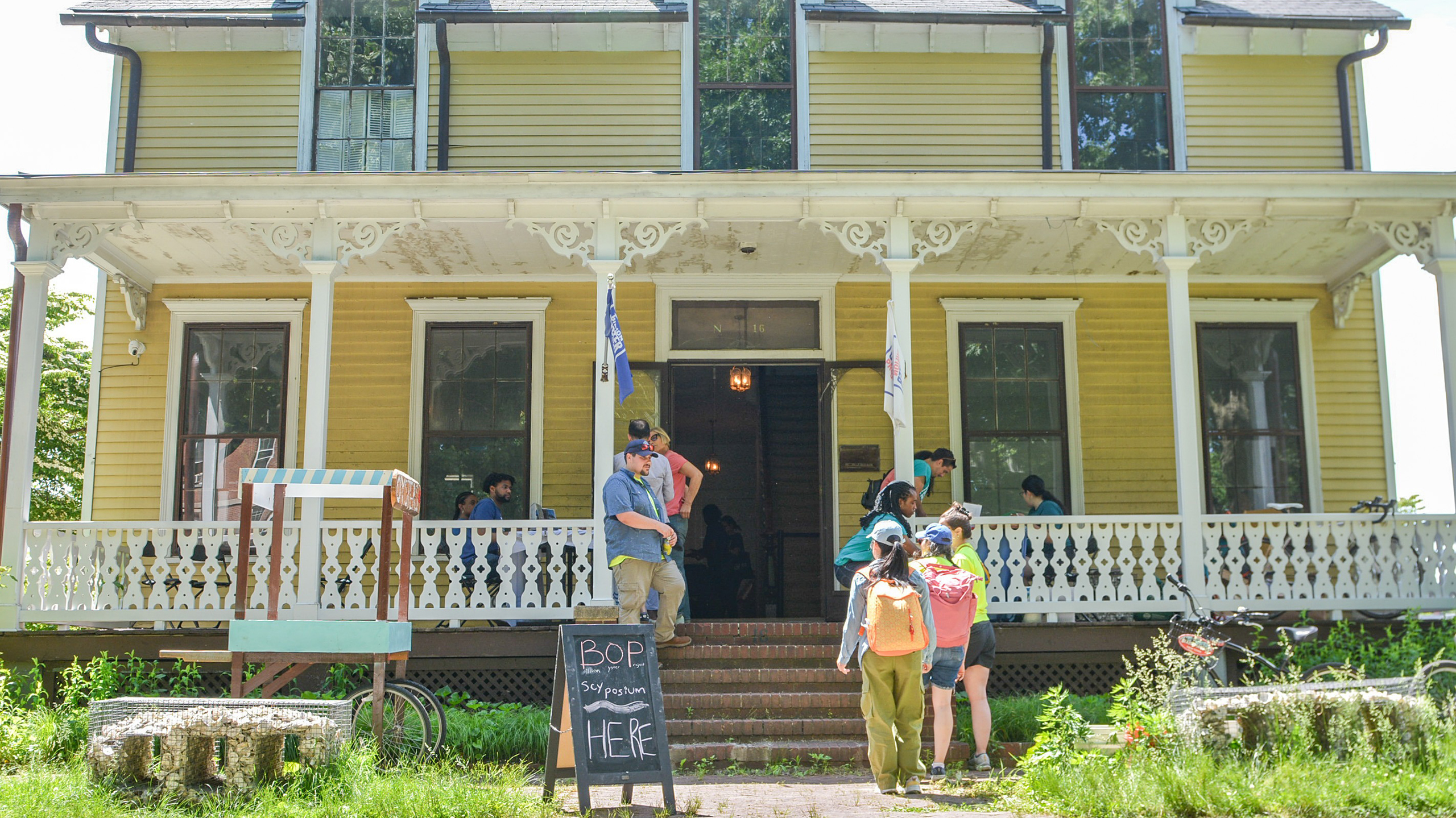 People gather on the porch of a large yellow house with a sign reading BOP shop open HERE out front.