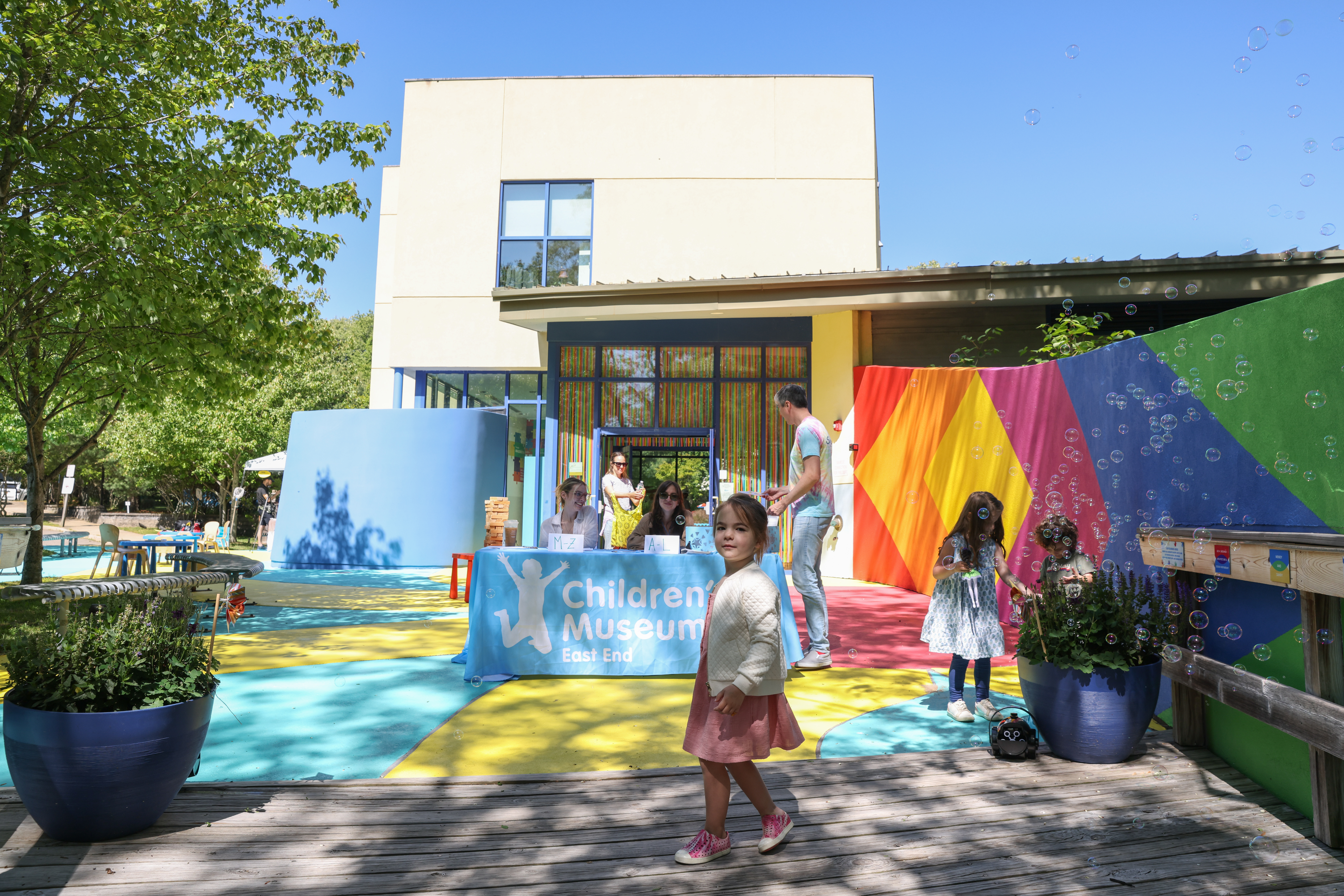 Children play with bubbles outside a colorful children’s museum on a sunny day.