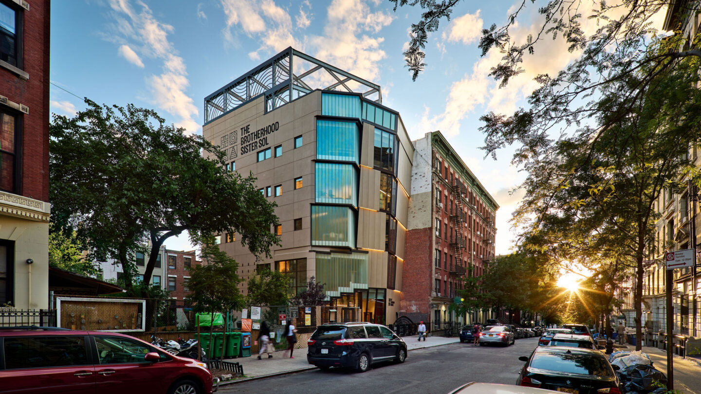 Modern building with glass and metal facade on a city street at sunset, with cars and people nearby.