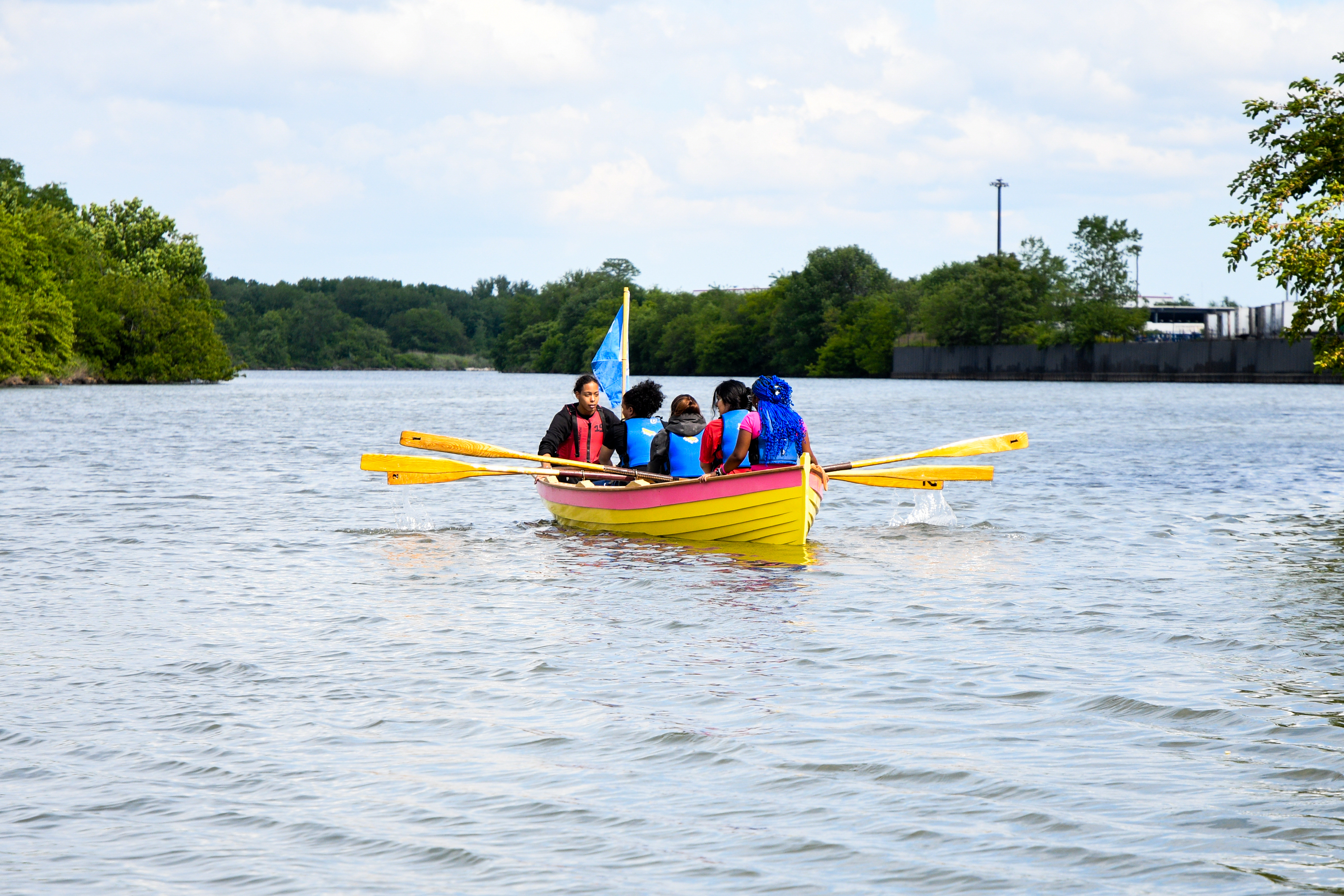 Person in a red life vest pushes a boat with rowers away from a dock over calm water.
