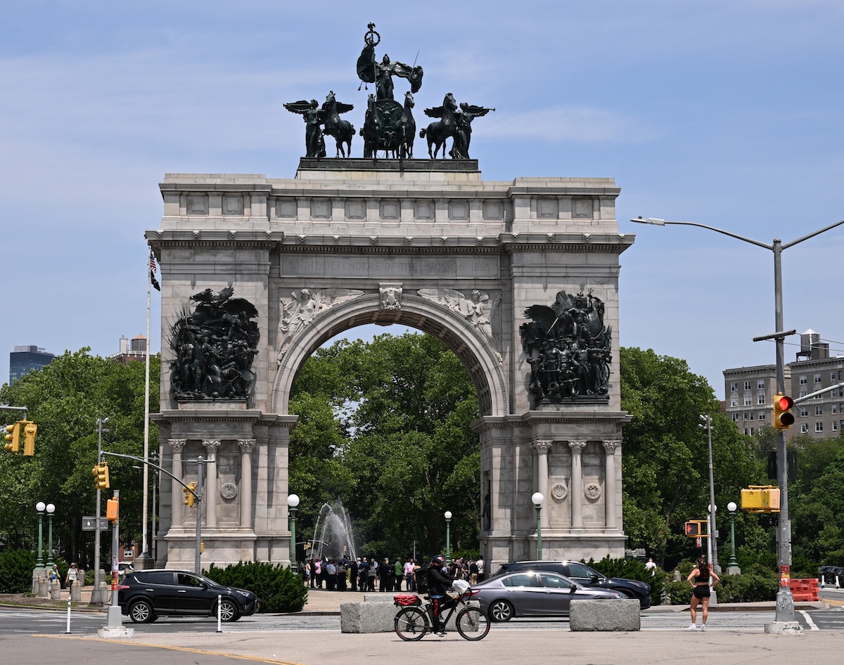 Grand Army Plaza’s stone triumphal arch with statues, people walking, and cyclists passing in Brooklyn, New York.