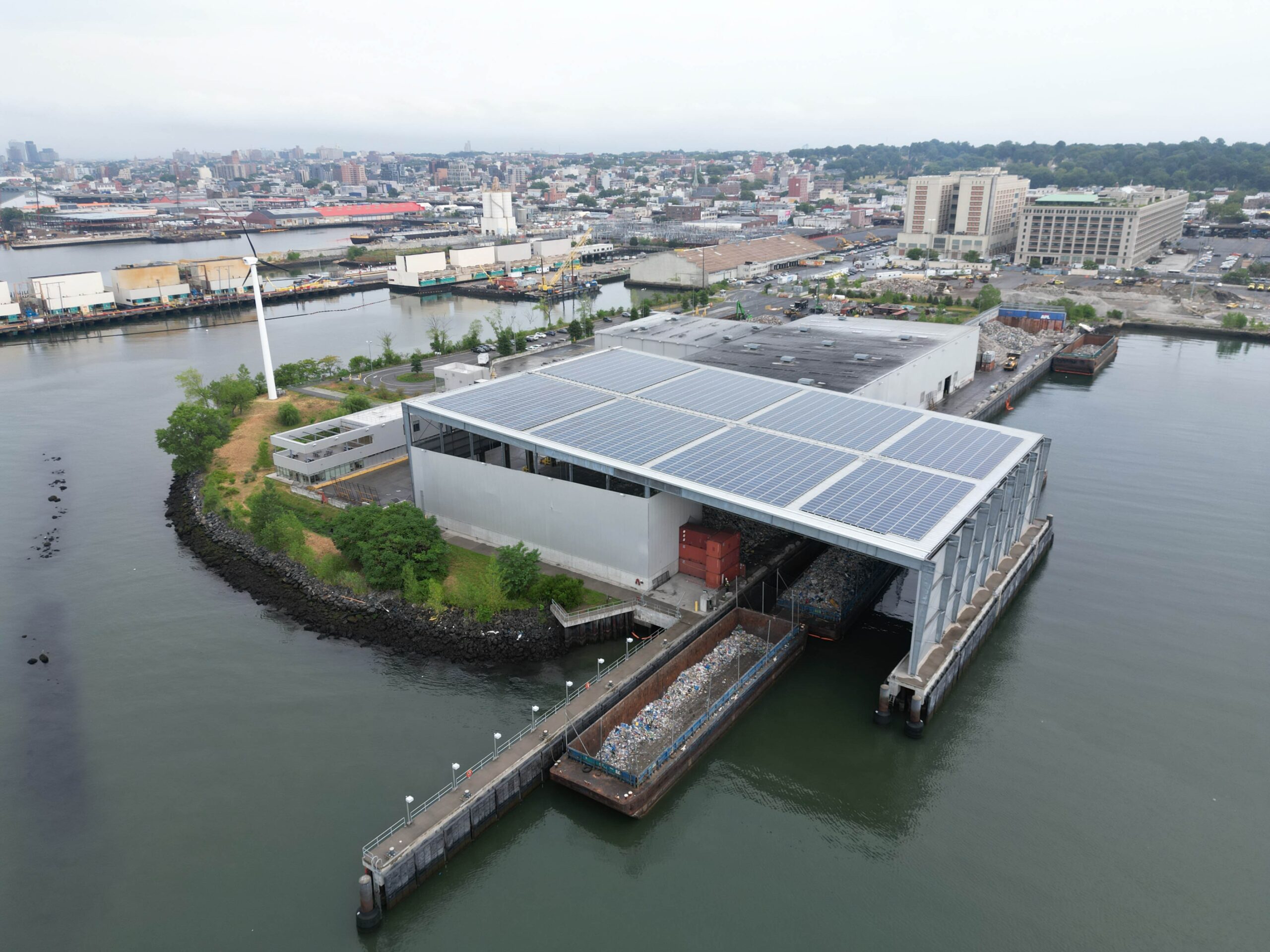 A waterfront industrial facility with a solar-panel roof, surrounded by water and city buildings in the background.
