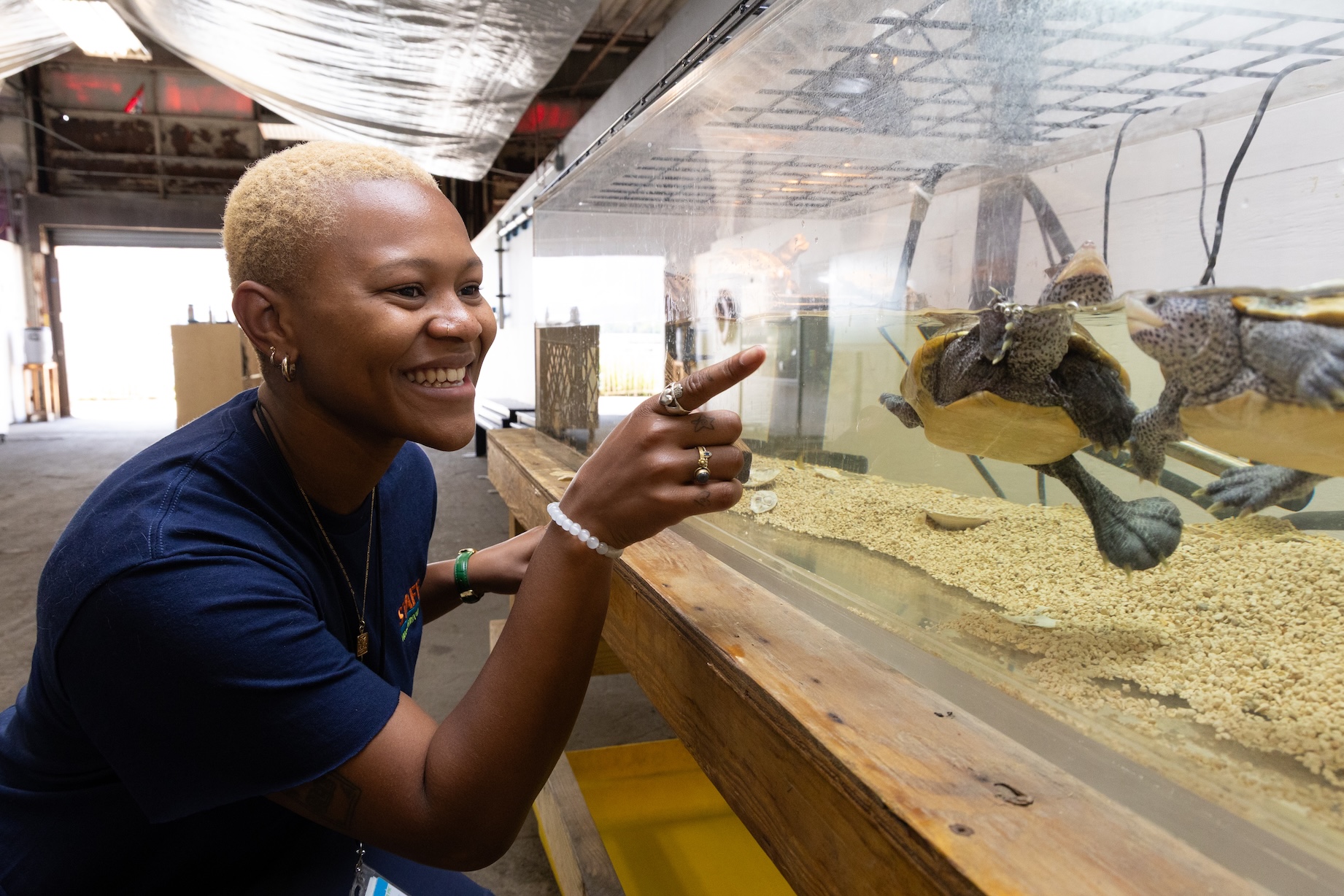 A person smiles and points at turtles swimming in a large aquarium inside a building.