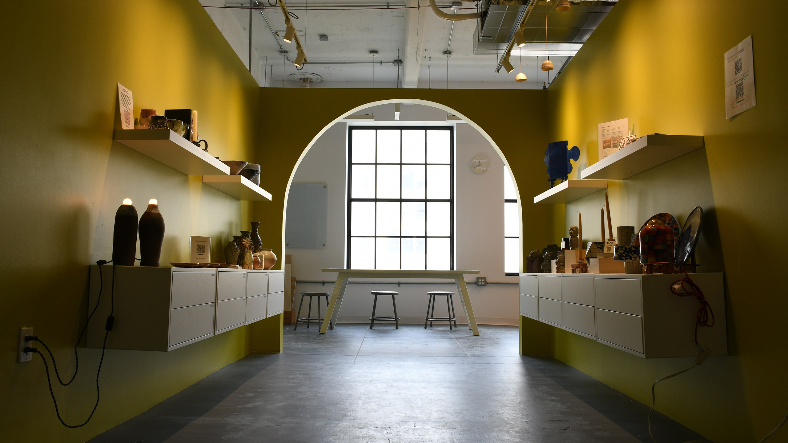 A modern gallery with yellow-green walls, shelves of pottery, and a large window with stools in the background.