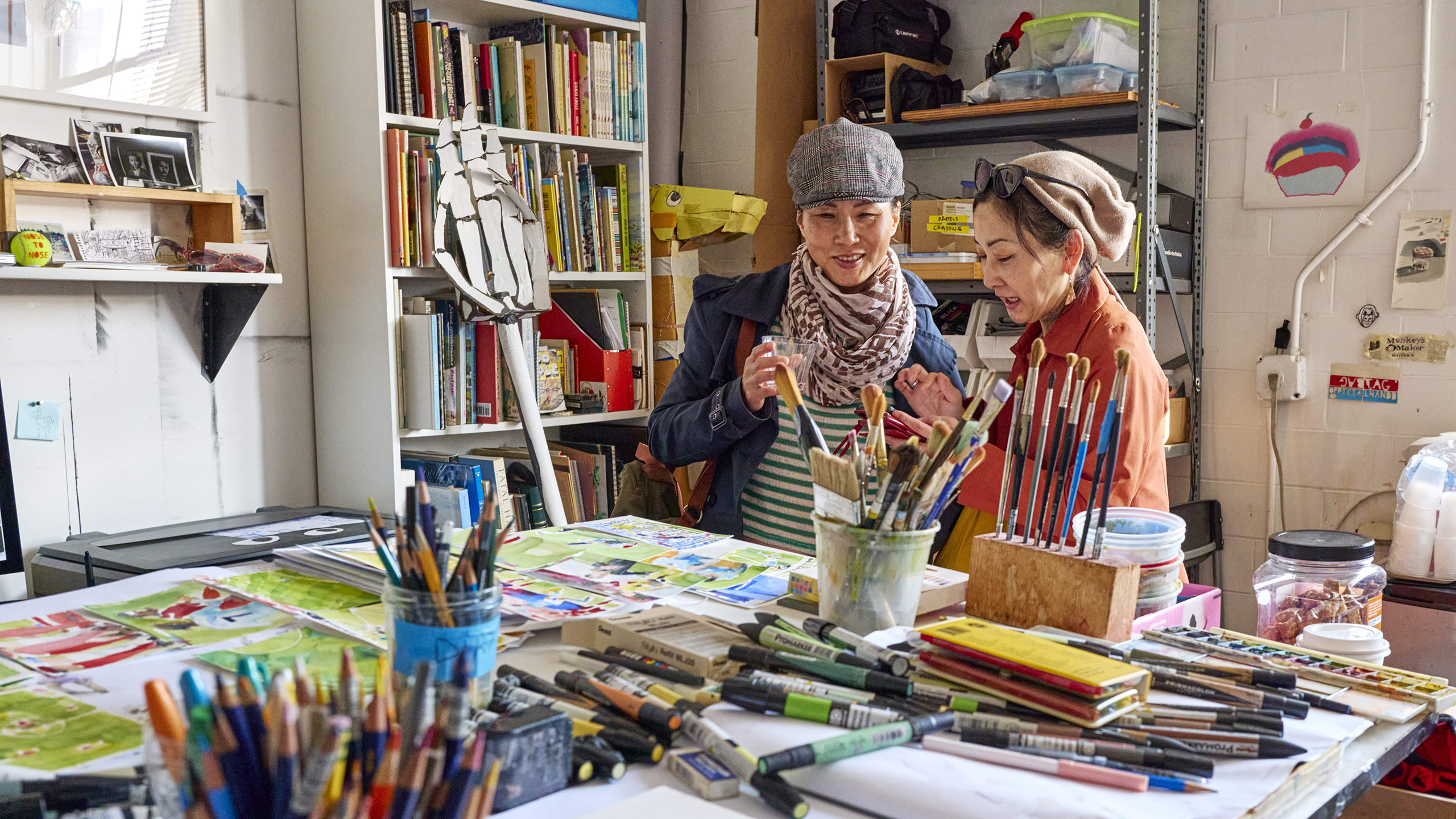 Two women talk in a bright art studio filled with books, art supplies, and colorful paintings.