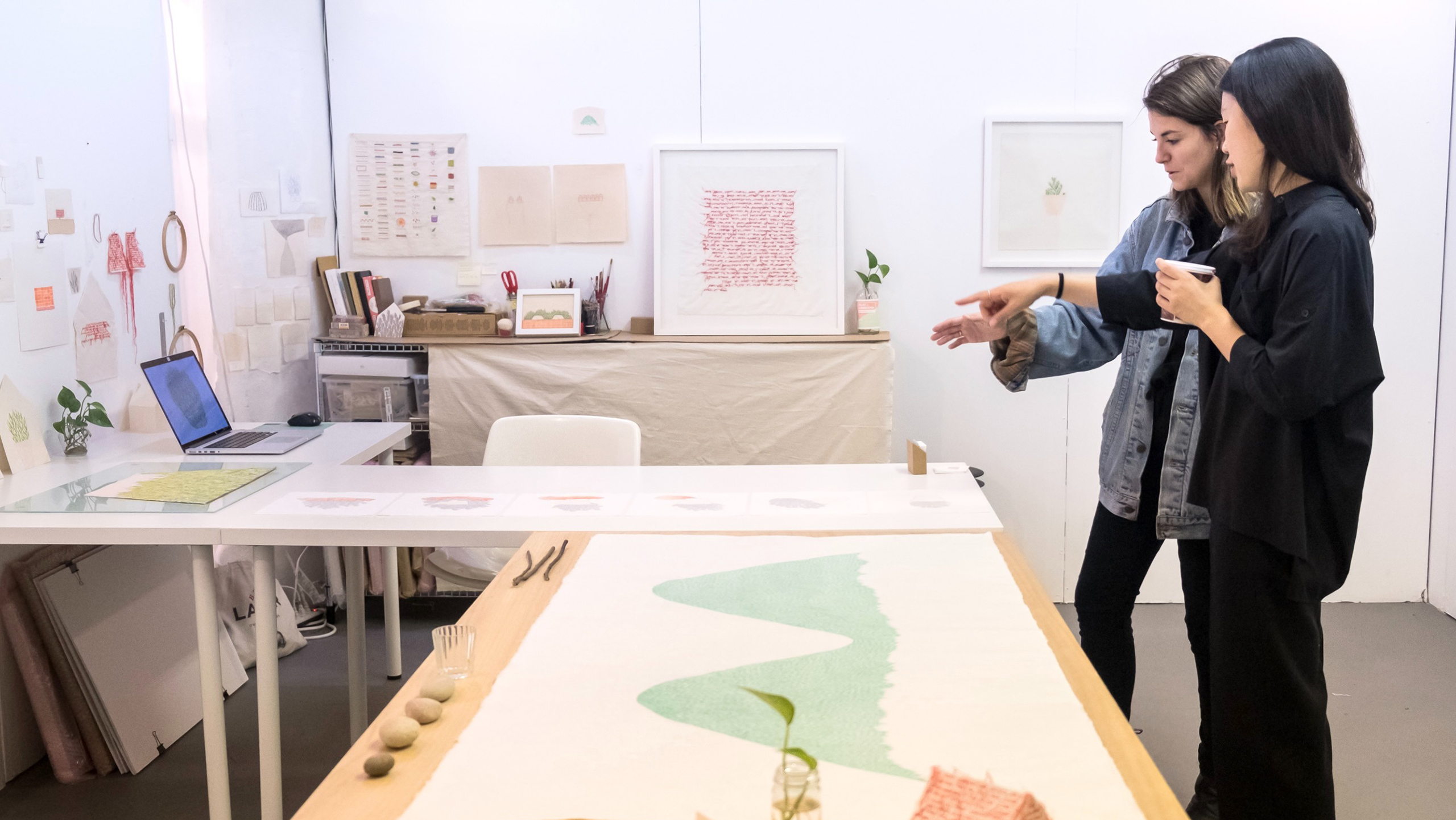 Two women discuss artwork in a bright studio with art supplies, prints, and a long illustrated cloth on a table.