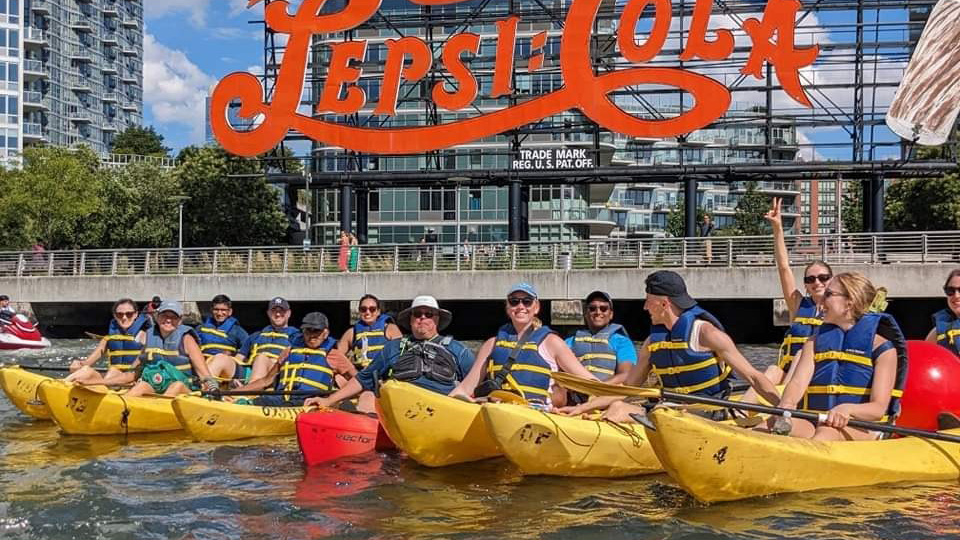 A group of people in yellow kayaks smile in front of a large red Pepsi-Cola sign by the water.