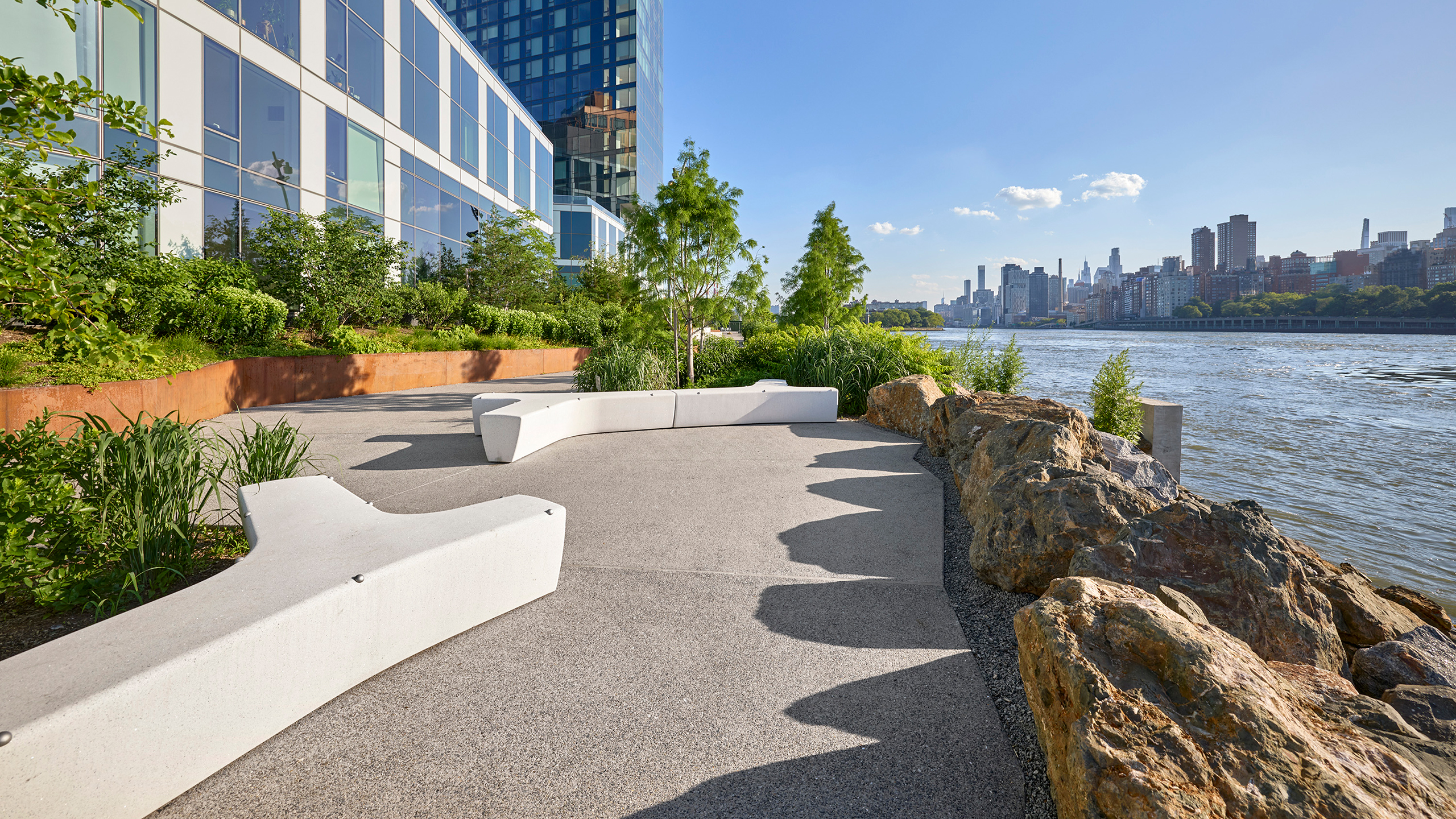 Curved white benches and greenery along a riverside path with city buildings in the background.