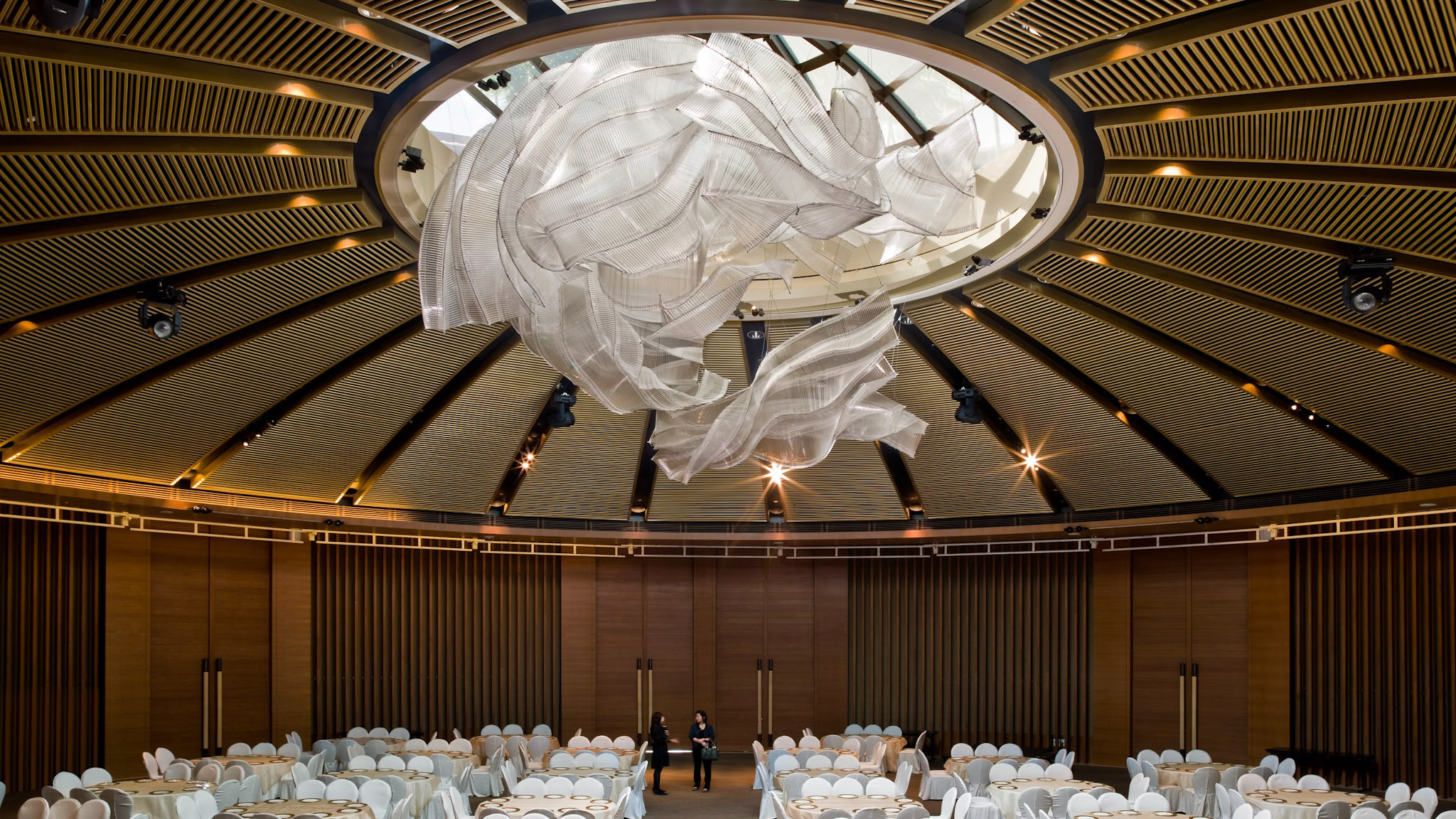 Round banquet hall with a dramatic ceiling sculpture and tables covered in white cloths; two people stand inside.