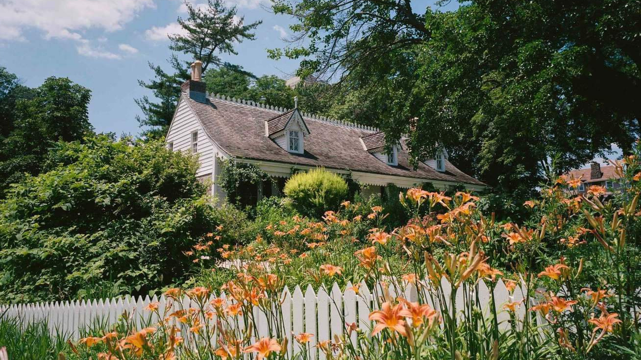 White cottage with steep roof behind a white picket fence and blooming orange lilies, surrounded by trees.