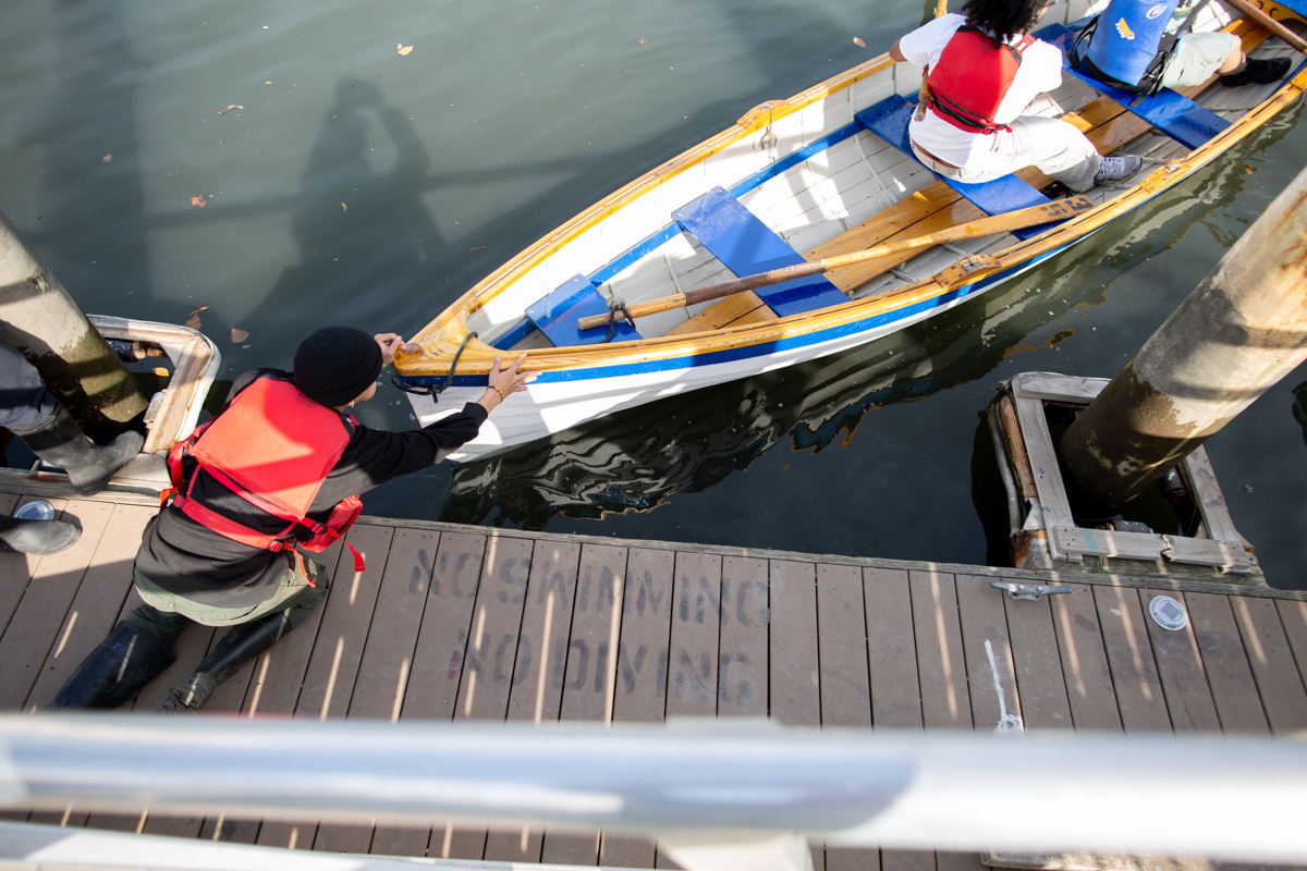 Person in a red life vest pushes a boat with rowers away from a dock over calm water.