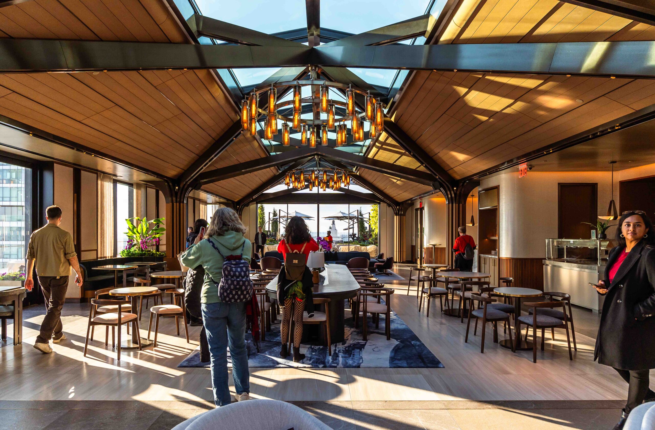 People walk and sit in a modern, sunlit lounge with wood decor and large windows revealing a city view.