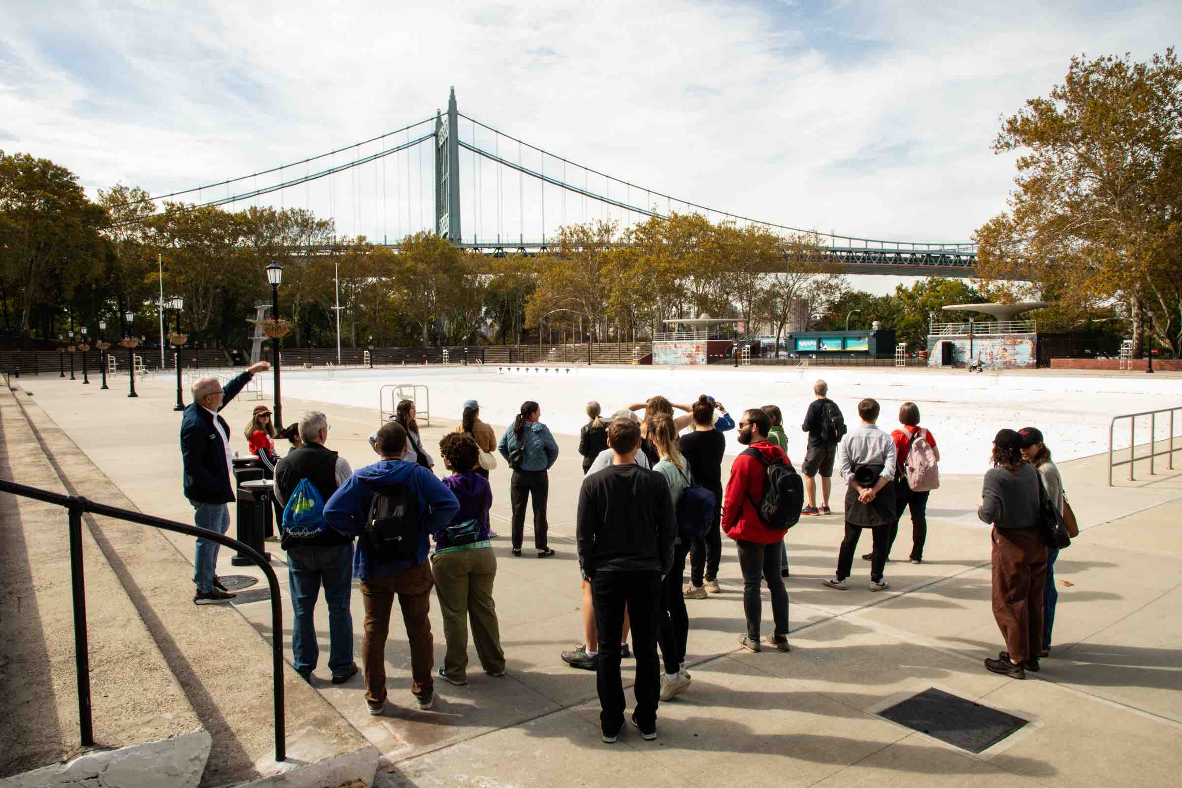 A group of people gather by an empty pool with a large suspension bridge in the background.