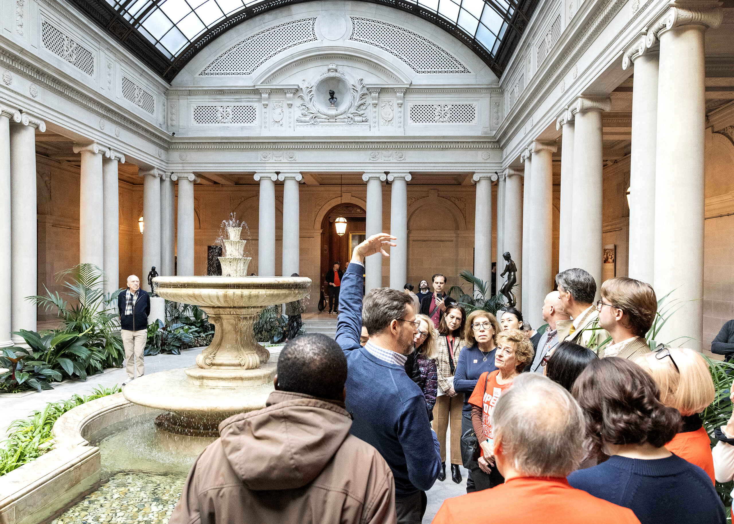 A tour guide gestures while speaking to a group in a grand hall with a fountain and classical columns.