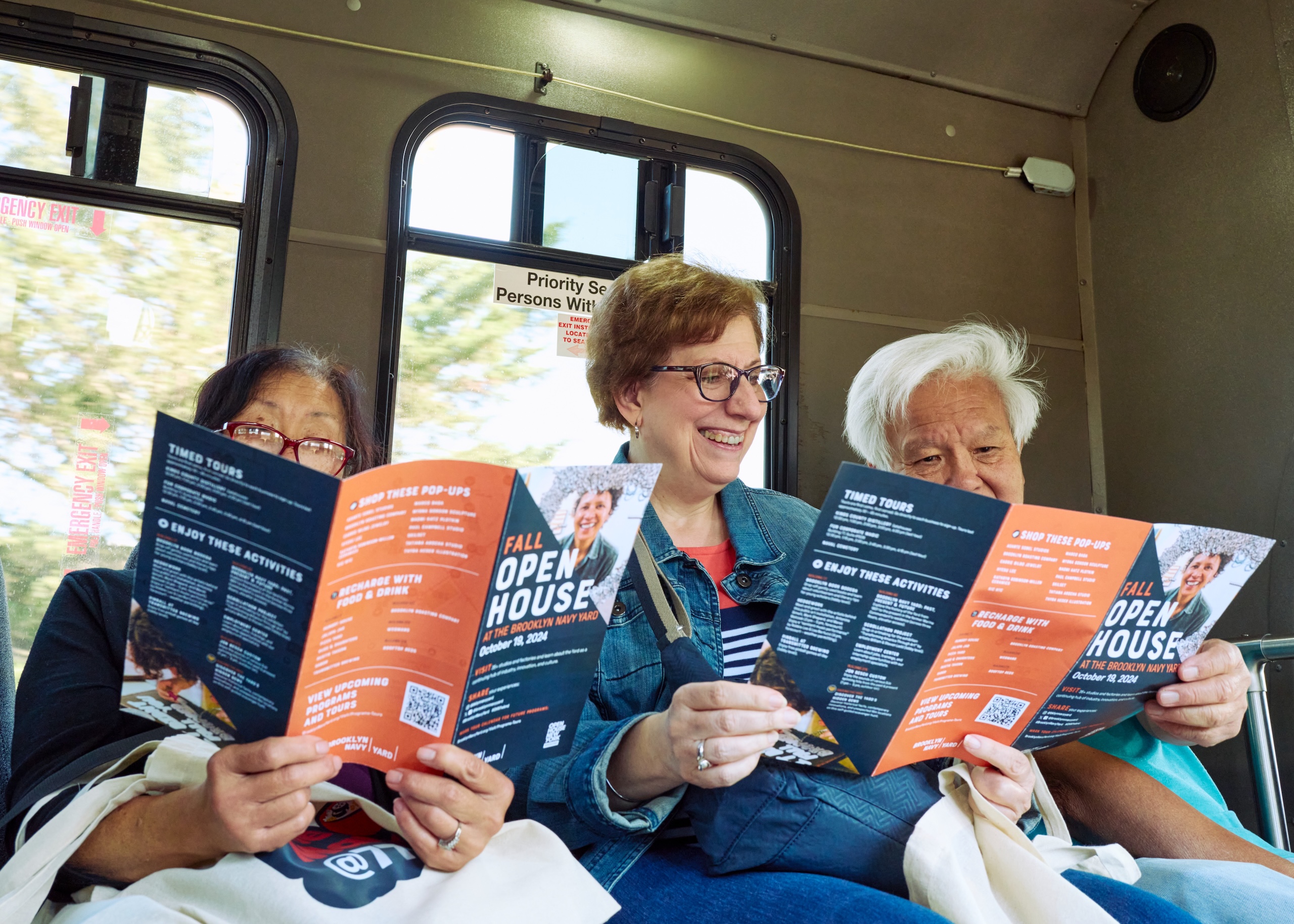 Three smiling people sitting on a bus, reading Fall Open House brochures together.