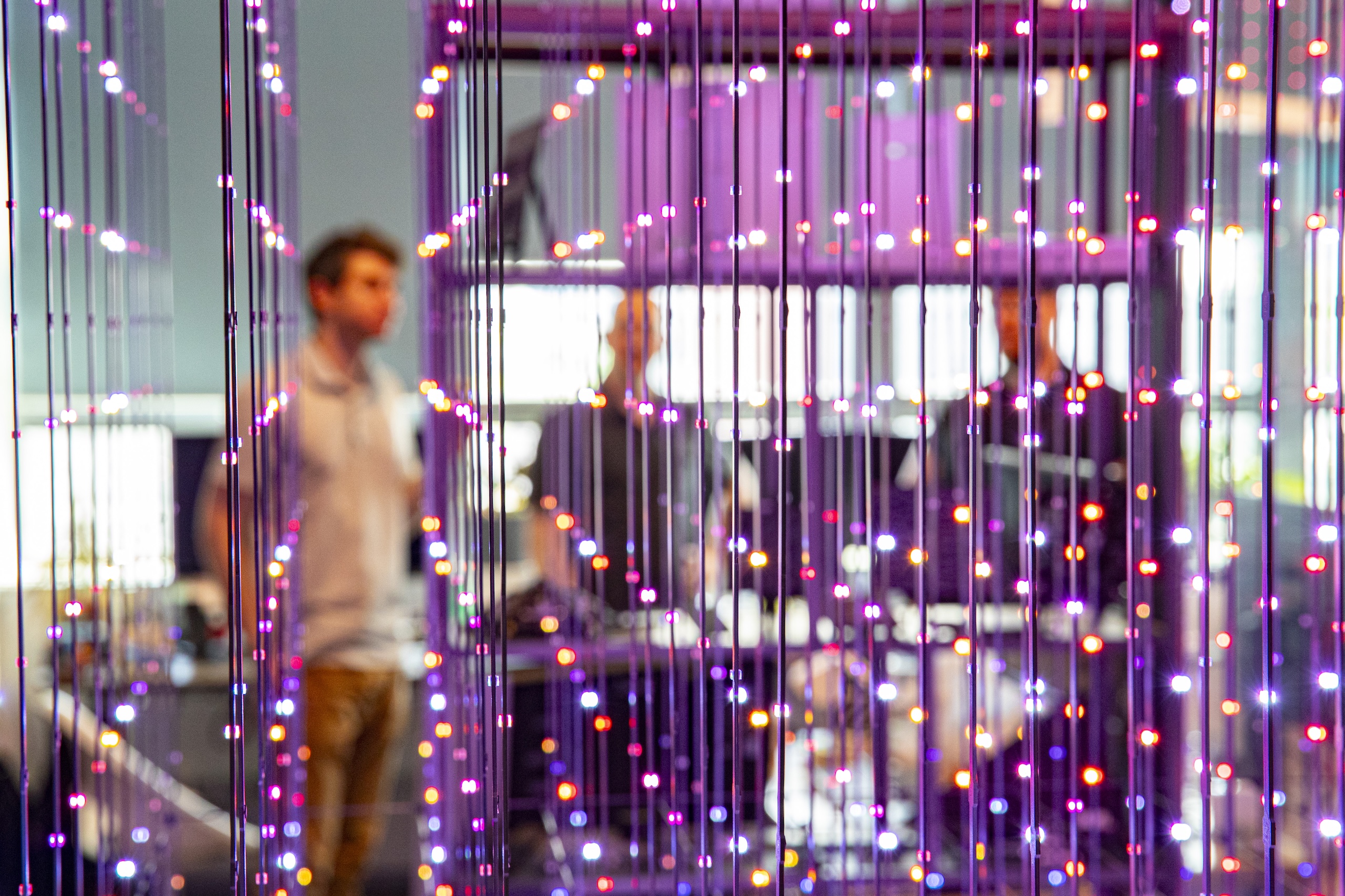 A man stands behind a curtain of colorful LED lights; two more blurred figures are visible in the background.