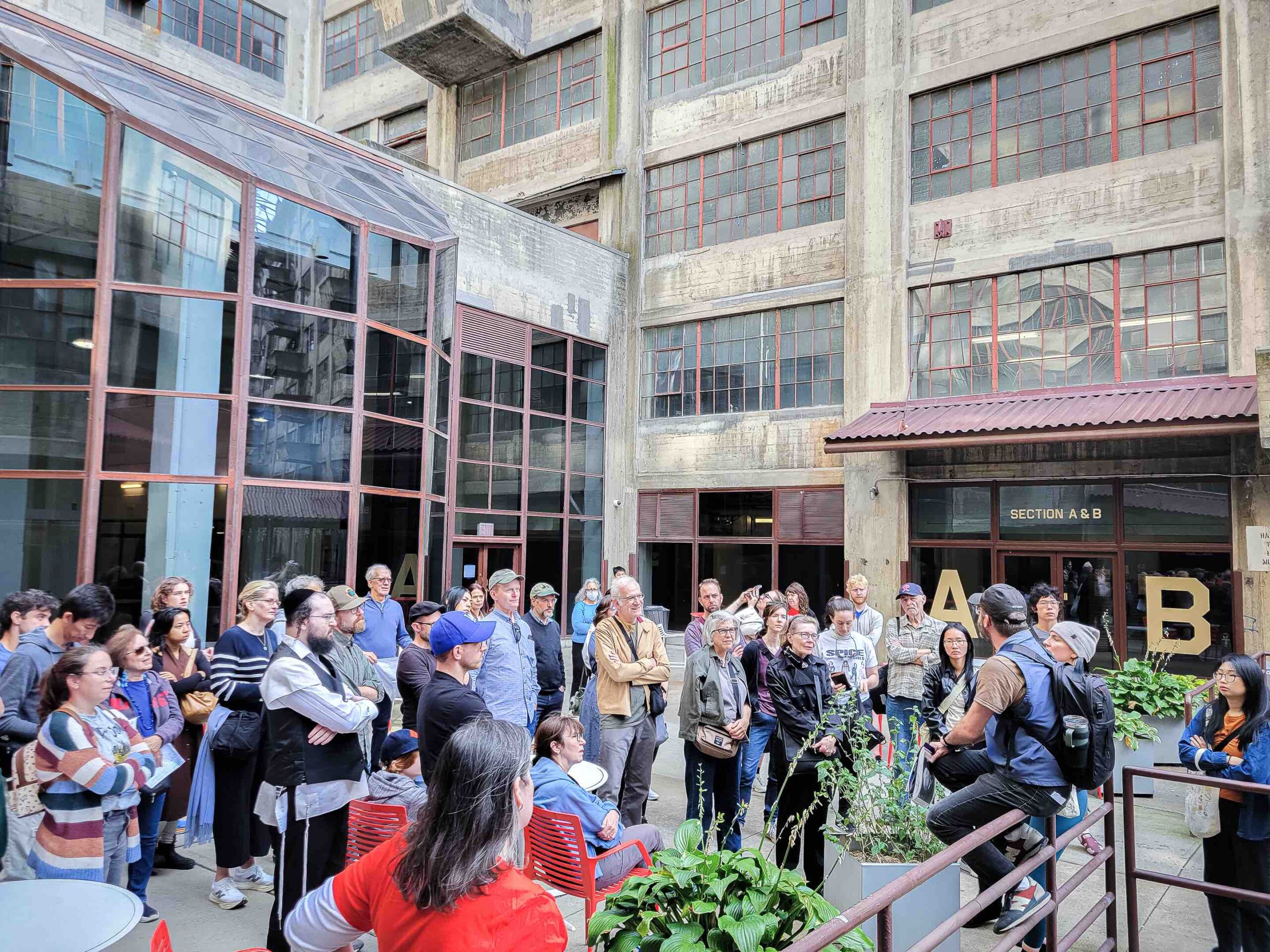 A group of people listen to a speaker in an urban courtyard surrounded by industrial-style buildings.