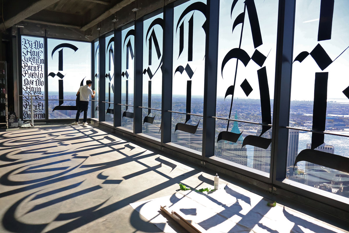 A person stands by tall windows with large black calligraphy casting shadows on the floor.