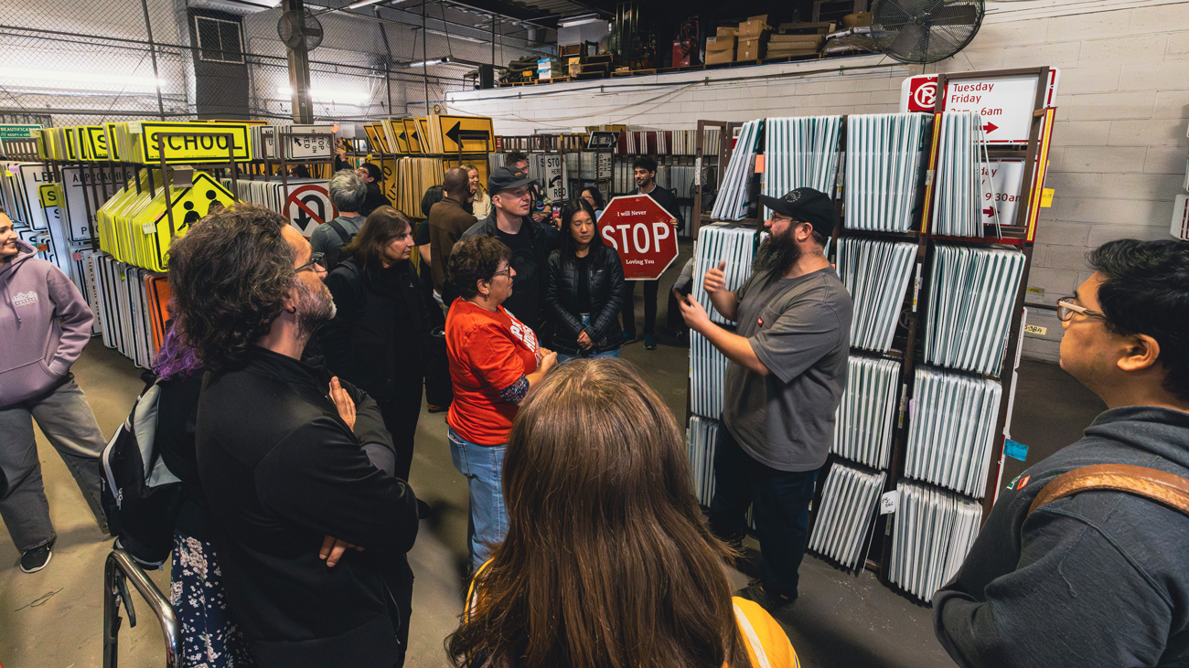 A group tours a sign warehouse as a guide explains traffic signs held up for the group to see.