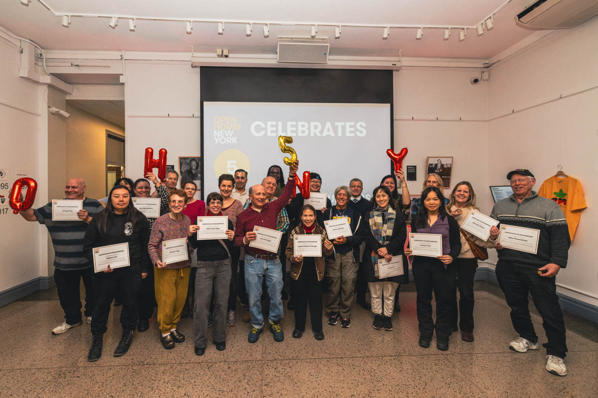 A group of people holding certificates stands in front of a screen that reads “CELEBRATES” in a gallery room.
